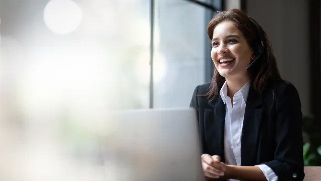 Smiling woman in a headset working on a laptop in a bright office environment.