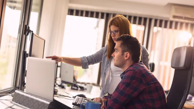 Woman and man collaborating at an office desk, focused on computer screen and discussing ideas.
