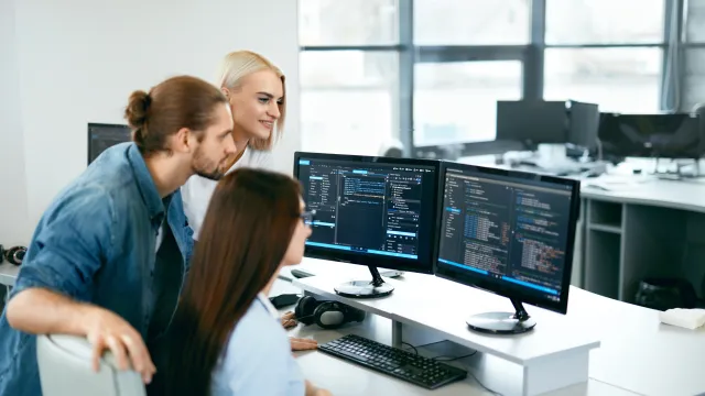 Three colleagues collaborating on programming tasks at their office desks with multiple monitors.
