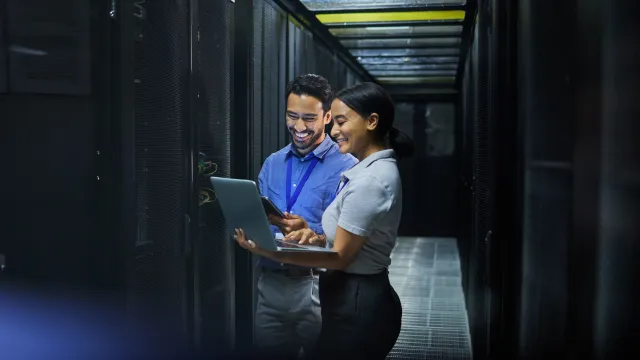 Two professionals collaborating in a data center, smiling while using a laptop and smartphone.