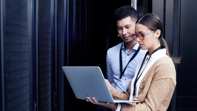 Two professionals collaborating on a laptop in a data center environment with server racks.