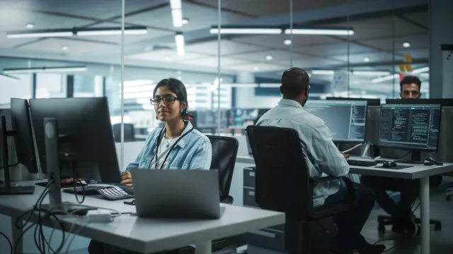 Team of diverse professionals collaborating in a modern office with computers and coding screens.