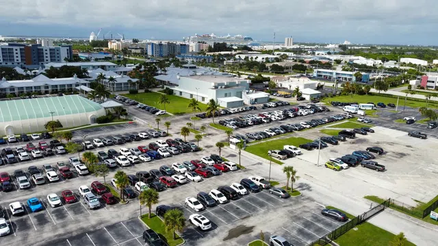 Aerial view of a large parking lot filled with cars near buildings and cruise ships in the distance under a cloudy sky.
