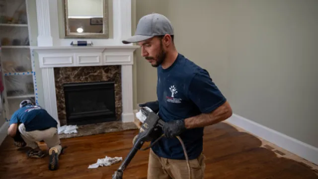 Two men sanding and refinishing hardwood floors in a living room with a fireplace and beige walls.