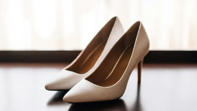 Pair of elegant beige high-heeled shoes with pointed toes on a wooden surface near a window.