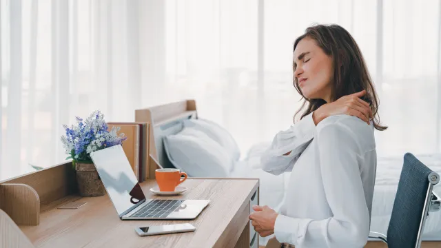 Young woman sitting at desk in bedroom rubbing her neck in discomfort with laptop and coffee cup on the table