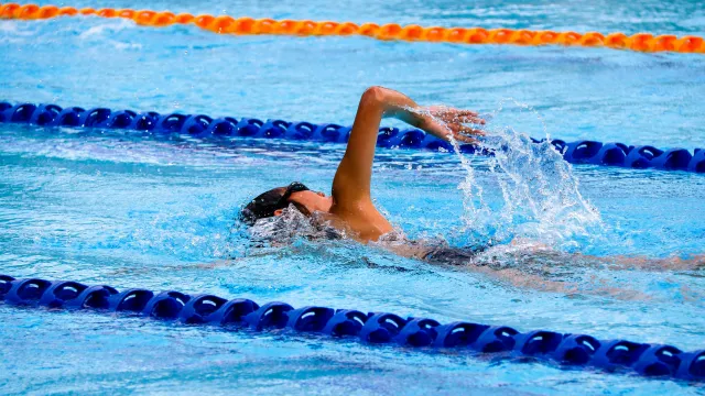 Swimmer performing backstroke in an outdoor pool with lane dividers and splashing water.