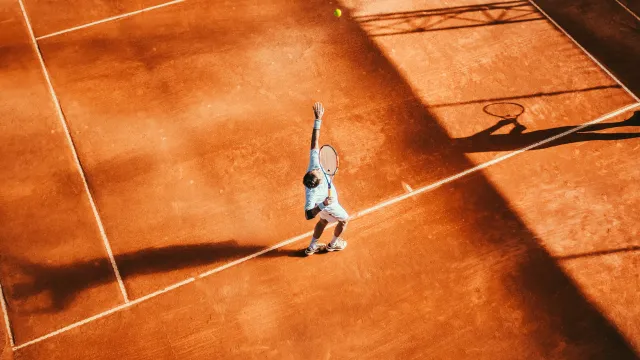 Tennis player serving ball on a sunlit clay court casting strong shadows during a match.