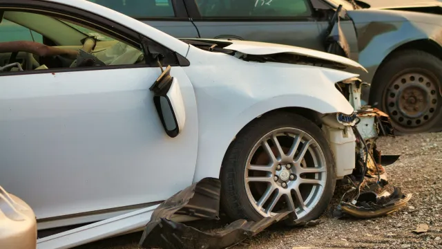White car with severe front-end damage and detached parts in a junkyard alongside other damaged vehicles.