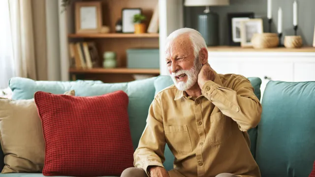 Elderly man sitting on couch holding neck in pain, surrounded by colorful pillows in a cozy living room.