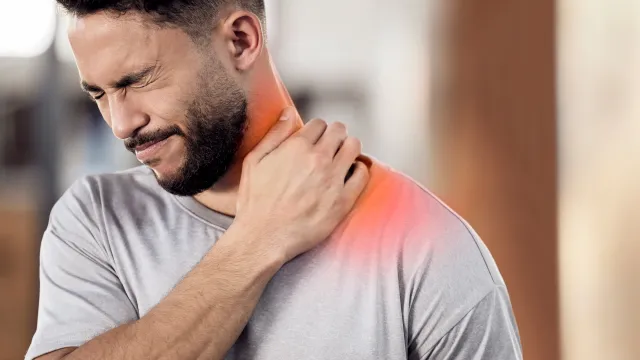 Man in gray shirt grimacing with hand on painful red shoulder and neck area indoors