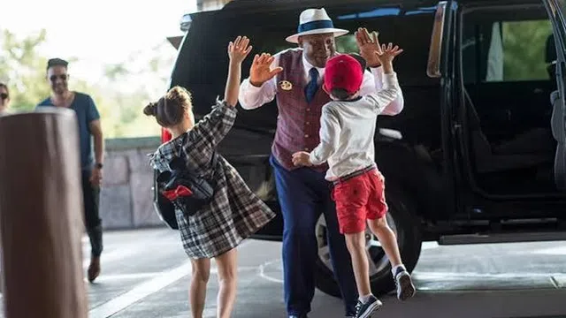 Hotel doorman greets two excited children arriving by black SUV with high fives outside bright entrance.