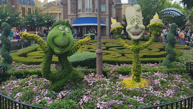 Topiary sculptures of animated characters in a flower garden with a central clock and historic buildings under a blue sky.