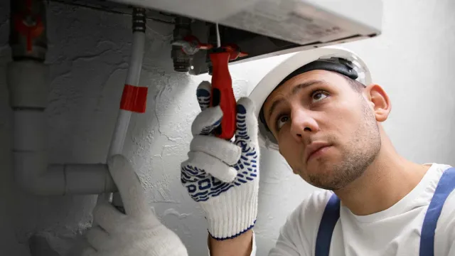 Plumber in white helmet and gloves fixing pipes under sink using a red wrench in a gray room.