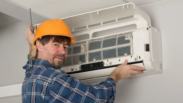 Technician in orange helmet repairing or installing wall-mounted air conditioner unit indoors.