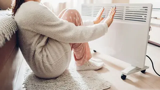 Woman in cozy clothes warming hands by a white electric heater in a bright living room.