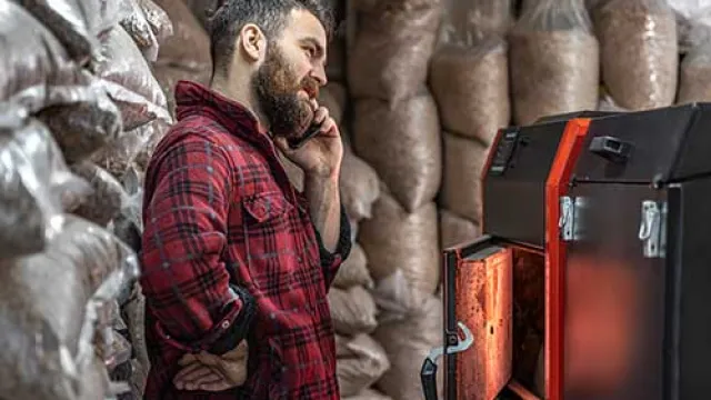 Man in red plaid shirt talking on phone next to a furnace with bags of pellets stacked behind him
