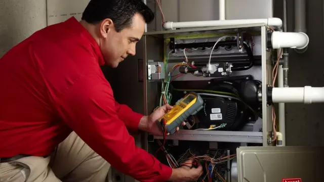Technician in red shirt diagnosing furnace system with multimeter inside an open Bryant HVAC unit.