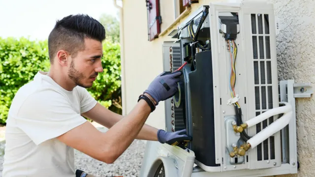 Technician repairing an outdoor air conditioning unit mounted on a wall using tools and wearing gloves.