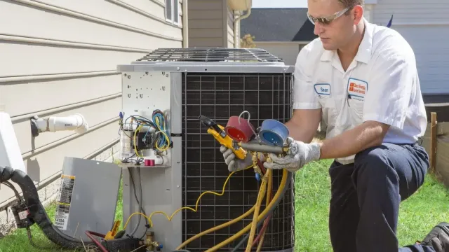 Technician in uniform servicing an outdoor air conditioning unit with gauges and tools on a residential lawn.