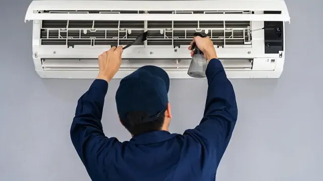 Technician cleaning and servicing a wall-mounted air conditioner with spray bottle and brush.