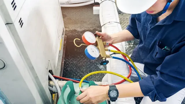 Technician in safety helmet testing air conditioning unit with pressure gauges and refrigerant tank outdoors.