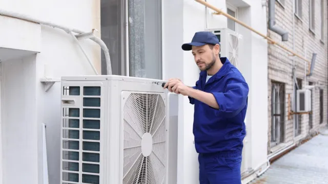 Technician in blue uniform repairing outdoor air conditioning unit on building exterior wall.