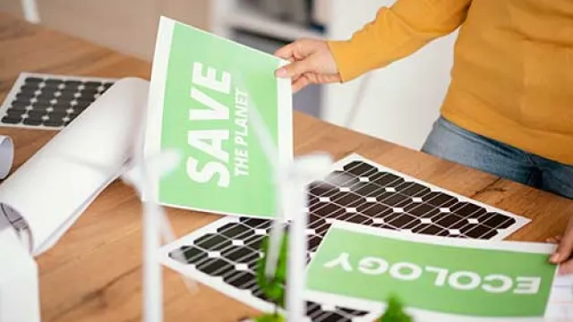 Person holding green eco-friendly posters saying Save the Planet and Ecology near solar panels and wind turbine models.