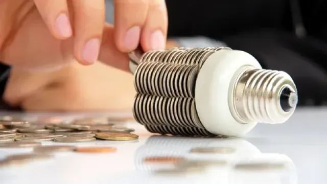 Hand placing coins near a light bulb made of stacked coins symbolizing energy savings and financial growth.