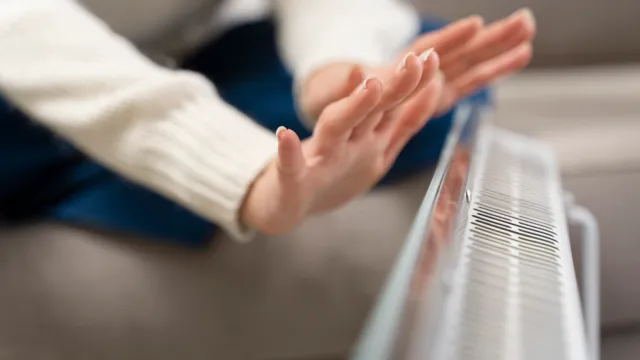 Person warming hands near a white electric heater in a cozy indoor setting.