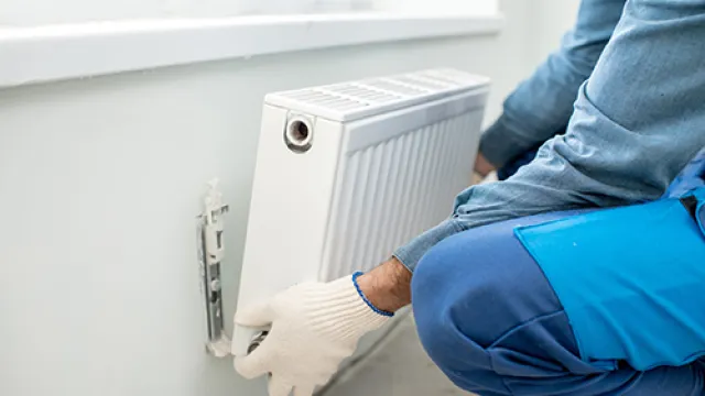 Technician in blue overalls installing a white wall-mounted radiator near a window indoors.