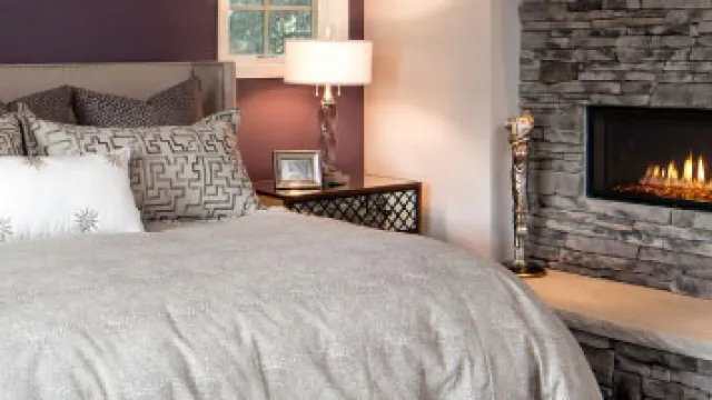 Cozy bedroom with a neatly made bed, decorative pillows, bedside lamp, and a modern stone fireplace.