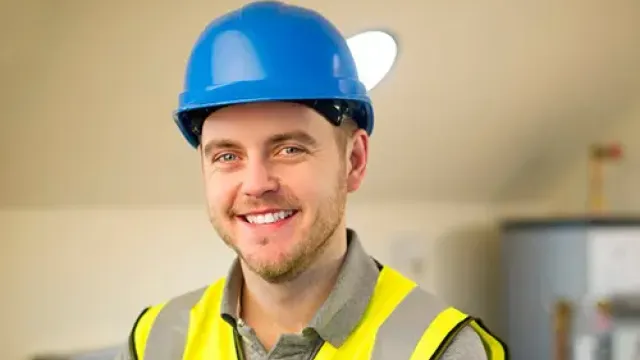 Smiling construction worker in blue hard hat and yellow safety vest standing with arms crossed indoors.