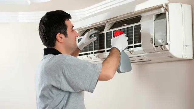 Technician wearing gloves cleaning and servicing a wall-mounted air conditioner indoors with a spray bottle.
