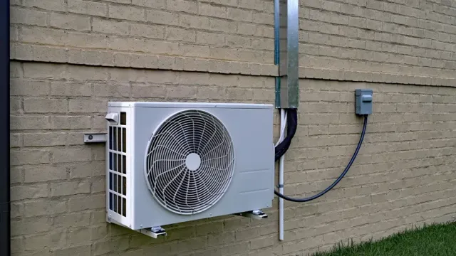 White outdoor air conditioning unit mounted on beige brick wall with green grass below and electrical box to the side