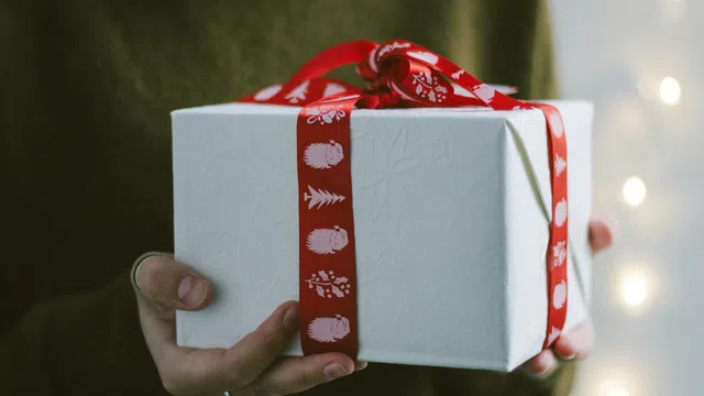 Person holding white gift box wrapped with red ribbon featuring holiday icons and bow on top.