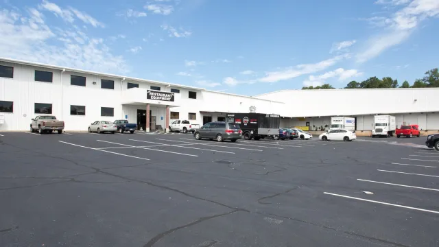 Large white industrial building with multiple parked vehicles and clear blue sky above in spacious parking lot.