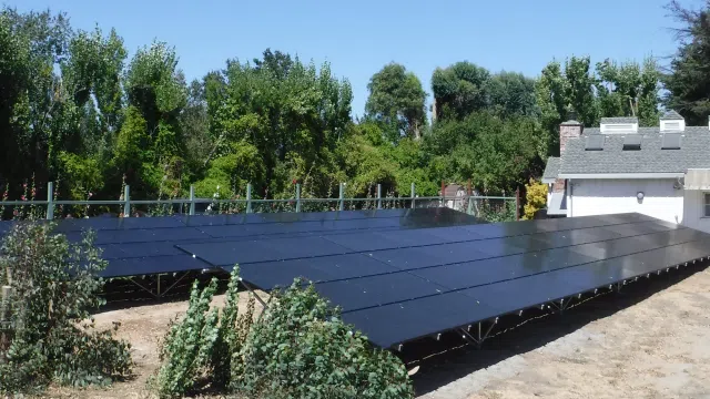 Large ground-mounted solar panel array beside a house with trees in the background on a sunny day