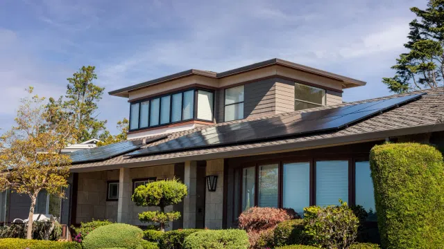 Modern house with solar panels on roof surrounded by manicured shrubs and trees under blue sky.