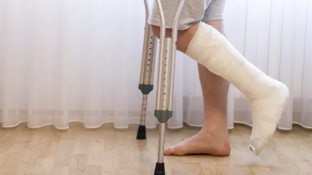 Person with a cast on foot using crutches standing on wooden floor in front of sheer curtains