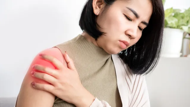 Woman with short black hair wearing sleeveless top holding painful red highlighted shoulder indoors.