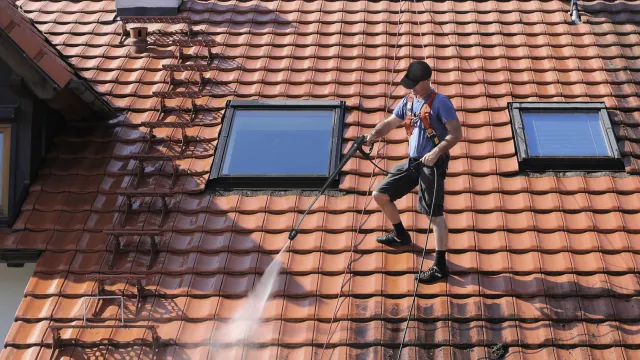Worker pressure washing dirt off a tiled roof while secured with a safety harness on a sunny day.