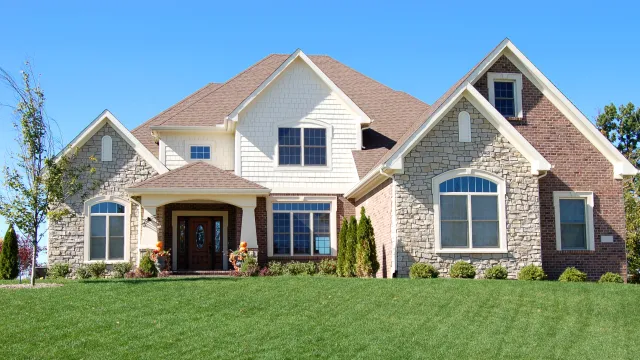 Large two-story suburban house with stone and brick exterior, manicured lawn, and clear blue sky background