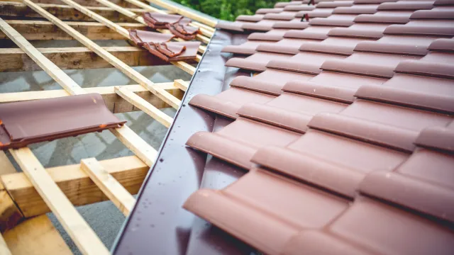Close-up of brown roof tiles being installed on wooden roof frame under construction with green background.