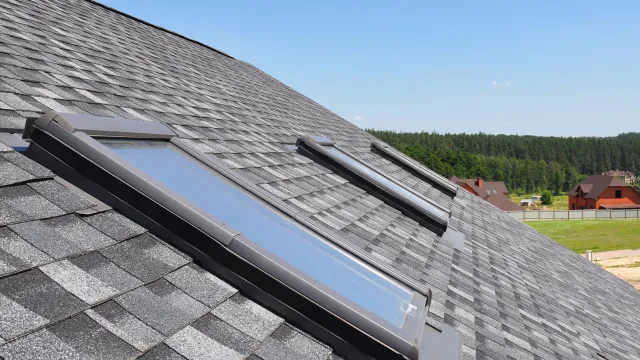 Close-up of roof with dark gray shingles and two skylight windows under clear blue sky.