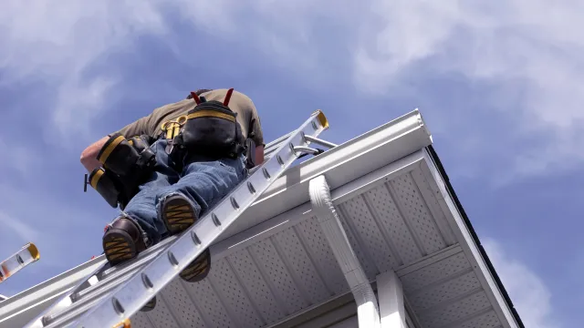 Worker climbing ladder to roof edge wearing tool belt against blue sky background