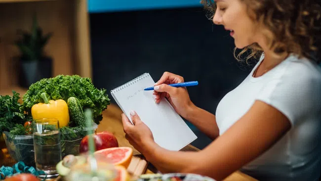 Woman writing on notepad near fresh vegetables and fruits on kitchen table for healthy meal planning