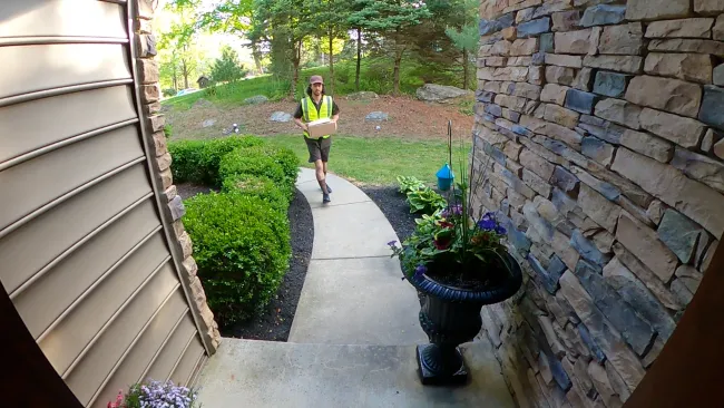 Delivery person walking on curved sidewalk towards house entrance holding a package in daylight.
