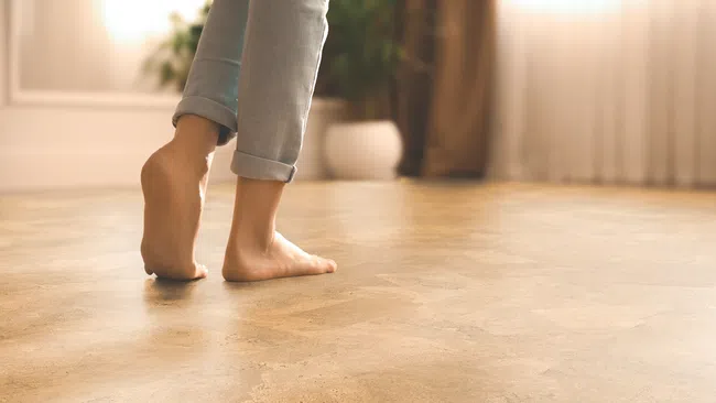 Close-up of bare feet on a warm-toned hardwood floor in a softly lit cozy room with indoor plants.
