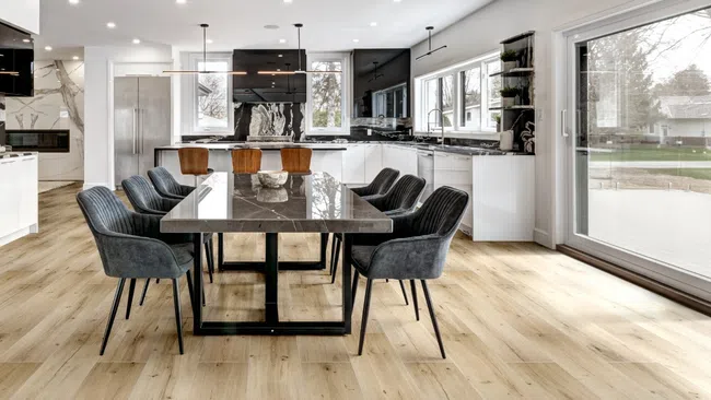 Modern kitchen dining area with a large marble table, gray upholstered chairs, wood flooring, and large windows.